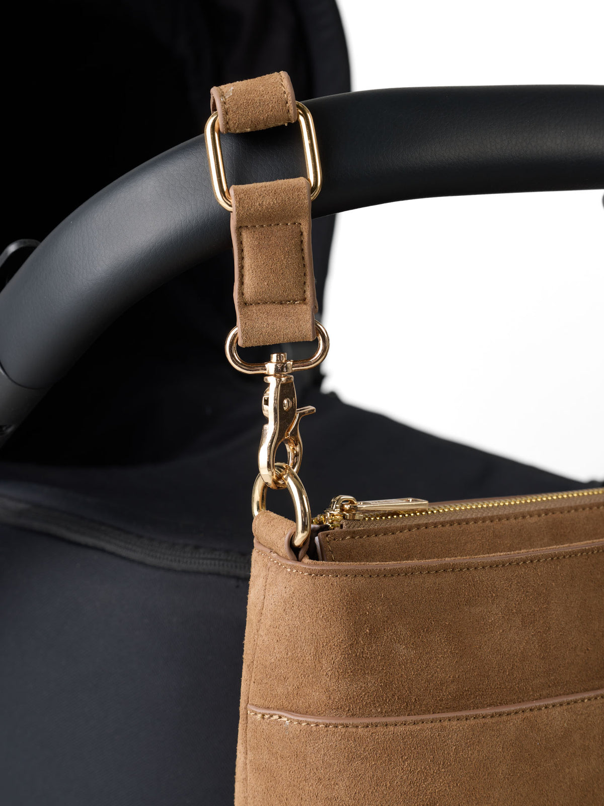 Close-up of a tan suede pram strap with gold-tone hardware attached to a black stroller handle, with part of a matching tan stroller bag visible - Sable Bebe