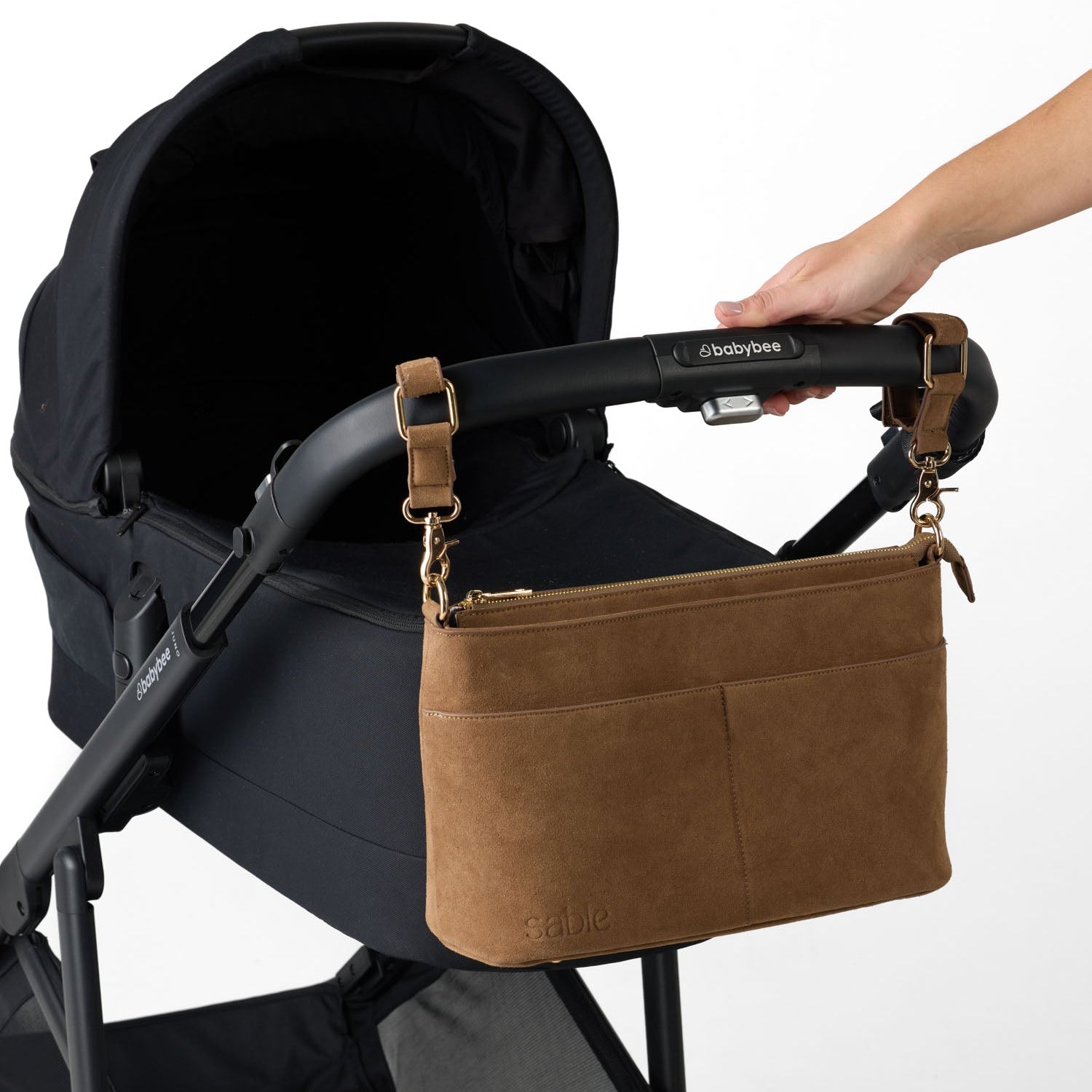 A brown suede baby shoulder bag with gold-tone hardware is shown hanging from the handle of a black stroller. A person's hand is holding the stroller handle to demonstrate the bag's placement - Sable Bebe