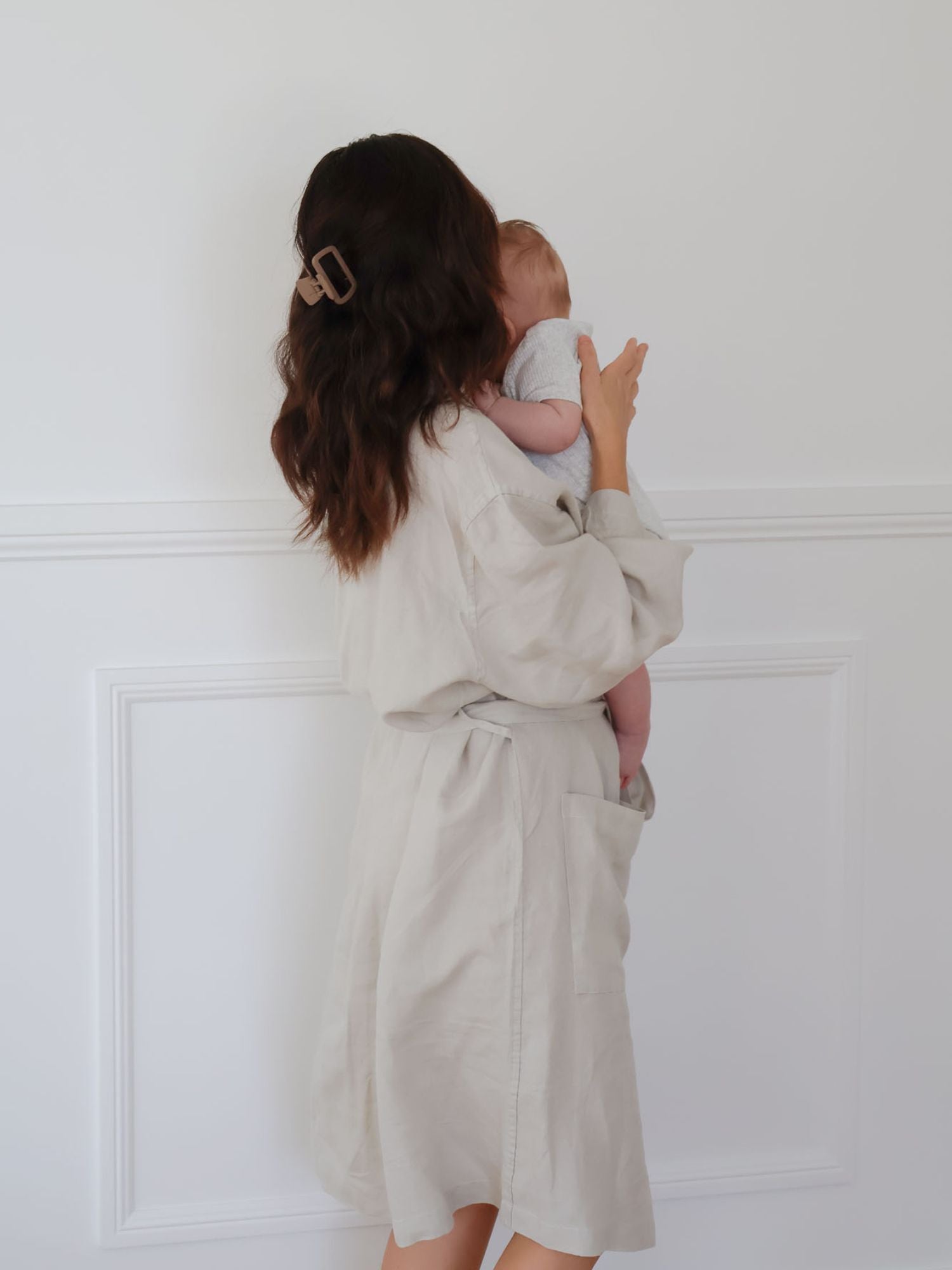 Woman in a beige robe holding a baby against a white wall
