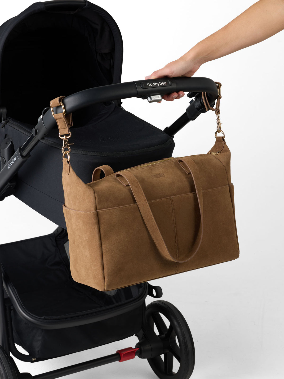 A brown suede-like diaper bag with gold-tone hardware is clipped to the handle of a black stroller. A person's arm is visible holding the stroller handle, demonstrating how the bag attaches.