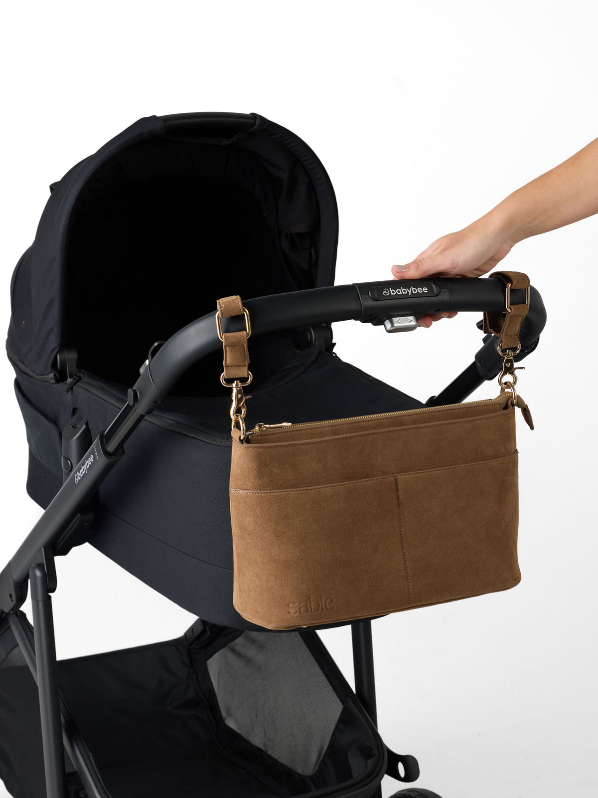 A brown suede baby shoulder bag with gold-tone hardware is shown hanging from the handle of a black stroller. A person's hand is holding the stroller handle to demonstrate the bag's placement - Sable Bebe