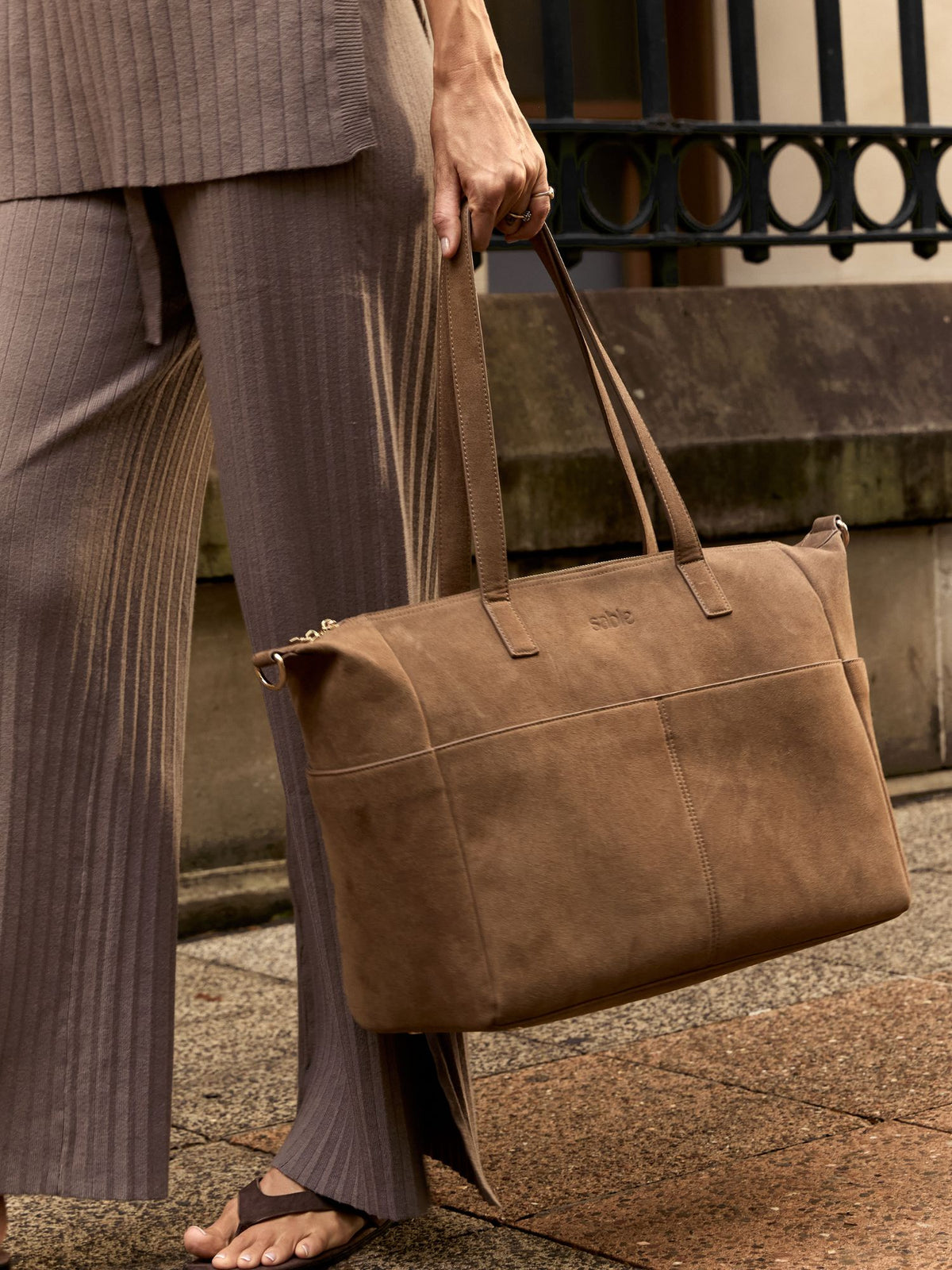 A person in taupe pleated trousers holds a large brown suede nappy bag with gold-tone hardware on a city sidewalk. - Sable Bebe